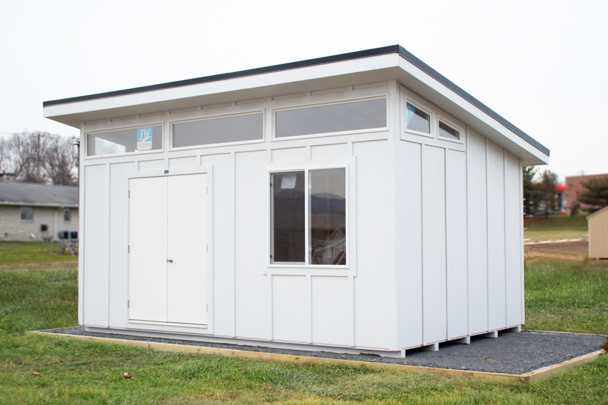 A white modern studio shed with a single-sloped roof, glass doors, and vertical board and batten siding. Positioned on a level gravel foundation with a dark walkway leading to the entrance.