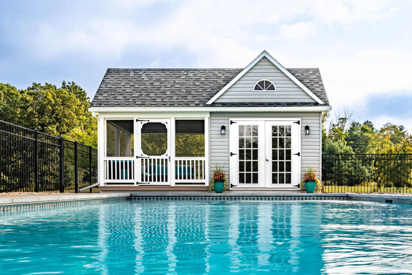 A charming prefab pool house with white siding, a gabled roof, and a screened porch sits adjacent to a large, crystal-clear in-ground swimming pool. The symmetrical design features double French doors with glass panes, flanked by two potted plants. The screened porch on the left side offers a shaded, enclosed space. The house has a gray shingled roof, a triangular vent near the peak, and a black fence enclosing the property. In the background, lush green trees and a partly cloudy sky provide a serene backdrop.